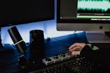 A person is operating a digital audio interface setup on a dark wooden desk. There is a microphone with a glowing LED ring, a mug, and a computer showcasing audio editing software with sound waves on the screen. The person’s hand is interacting with dials on an audio control interface.