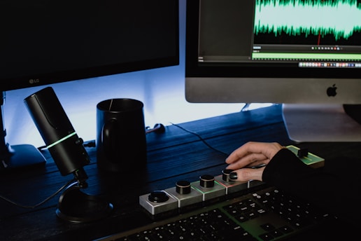 A person is operating a digital audio interface setup on a dark wooden desk. There is a microphone with a glowing LED ring, a mug, and a computer showcasing audio editing software with sound waves on the screen. The person’s hand is interacting with dials on an audio control interface.