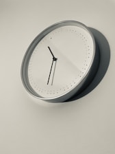 A minimalist wall clock with a silver frame is mounted on a plain wall. The clock face is white, featuring black hour and minute hands and small black dots marking the hours. The overall aesthetic is modern and simple.