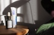 A wooden surface holds two skincare products: a small dark glass bottle with a dropper labeled 'U.F.O. Acne Treatment Face Oil' and a tube labeled 'Differin Gel.' Soft shadows and light create a calm, minimalistic setting with hints of foliage in the corner.