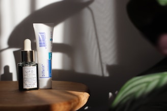 A wooden surface holds two skincare products: a small dark glass bottle with a dropper labeled 'U.F.O. Acne Treatment Face Oil' and a tube labeled 'Differin Gel.' Soft shadows and light create a calm, minimalistic setting with hints of foliage in the corner.