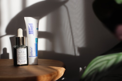 A wooden surface holds two skincare products: a small dark glass bottle with a dropper labeled 'U.F.O. Acne Treatment Face Oil' and a tube labeled 'Differin Gel.' Soft shadows and light create a calm, minimalistic setting with hints of foliage in the corner.
