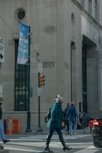 A city street scene features people crossing at a pedestrian crosswalk near a large building with the address 1500 Walnut visible. There are traffic signs, a streetlamp, and a large banner for a park. One person in a teal coat and mask is prominent in the foreground, while others walk in different directions. The atmosphere is urban and somewhat busy.