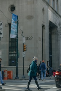 A city street scene features people crossing at a pedestrian crosswalk near a large building with the address 1500 Walnut visible. There are traffic signs, a streetlamp, and a large banner for a park. One person in a teal coat and mask is prominent in the foreground, while others walk in different directions. The atmosphere is urban and somewhat busy.