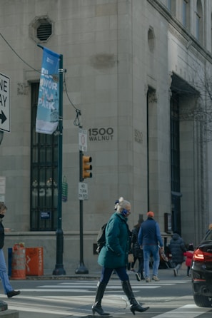 A city street scene features people crossing at a pedestrian crosswalk near a large building with the address 1500 Walnut visible. There are traffic signs, a streetlamp, and a large banner for a park. One person in a teal coat and mask is prominent in the foreground, while others walk in different directions. The atmosphere is urban and somewhat busy.