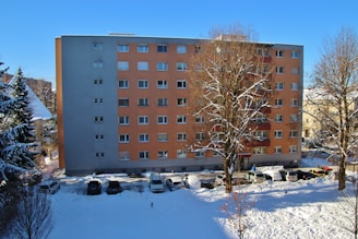 A residential apartment building with a flat facade, featuring light orange and gray colors. In front, several leafless trees partially covered with snow can be seen under a clear blue sky. A line of parked cars are visible along the snow-covered street at the building's base.