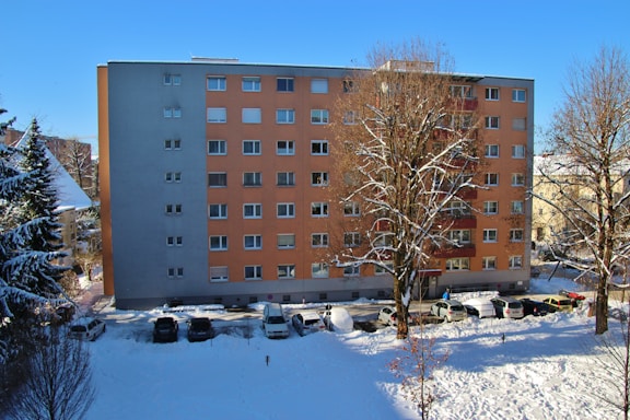 A residential apartment building with a flat facade, featuring light orange and gray colors. In front, several leafless trees partially covered with snow can be seen under a clear blue sky. A line of parked cars are visible along the snow-covered street at the building's base.