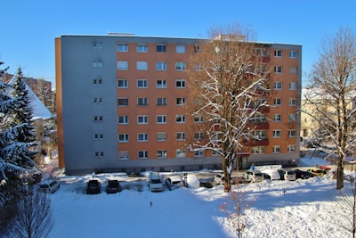 A residential apartment building with a flat facade, featuring light orange and gray colors. In front, several leafless trees partially covered with snow can be seen under a clear blue sky. A line of parked cars are visible along the snow-covered street at the building's base.