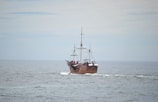 A vintage-style sailing ship with multiple masts is navigating through calm ocean waters under an overcast sky. The ship has a wooden appearance and features several white sails, though they are furled. People are visible on board, adding a sense of activity.