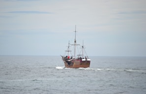 A vintage-style sailing ship with multiple masts is navigating through calm ocean waters under an overcast sky. The ship has a wooden appearance and features several white sails, though they are furled. People are visible on board, adding a sense of activity.