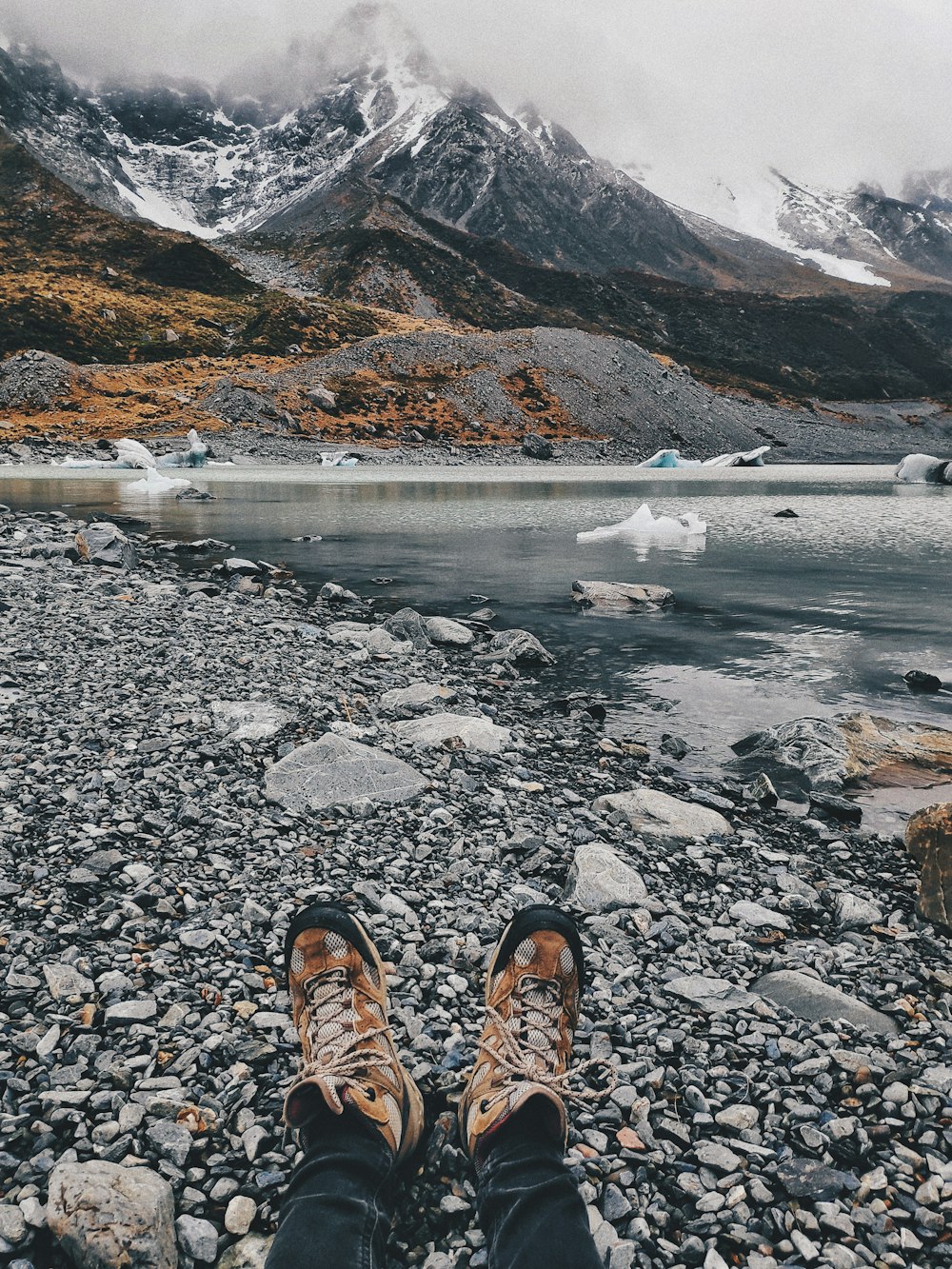 person in brown hiking shoes standing on rocky ground near body of water during daytime