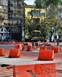 Barriers and orange mesh fencing are set up around a construction area on the street, with a yellow building and surrounding trees in the background. The barriers are placed in an orderly fashion, indicating ongoing roadwork or maintenance.