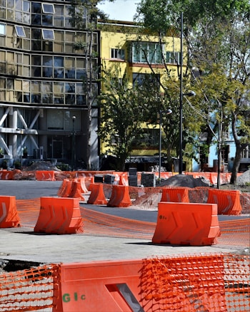Barriers and orange mesh fencing are set up around a construction area on the street, with a yellow building and surrounding trees in the background. The barriers are placed in an orderly fashion, indicating ongoing roadwork or maintenance.