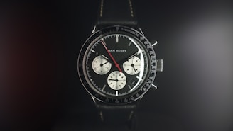 A detailed wristwatch with a black leather strap and a black bezel featuring white and red markers. The watch face displays three sub-dials, with the brand name visible on the face. The ambient lighting creates a soft glow around the watch, highlighting its polished features.