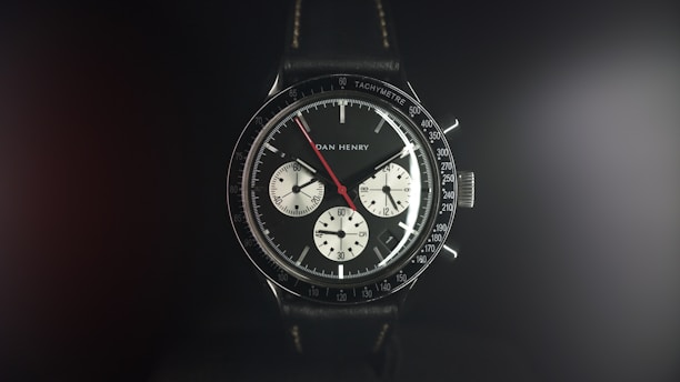 A detailed wristwatch with a black leather strap and a black bezel featuring white and red markers. The watch face displays three sub-dials, with the brand name visible on the face. The ambient lighting creates a soft glow around the watch, highlighting its polished features.