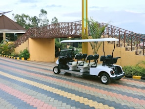 A white golf cart is parked on a multicolored brick-paved path. Behind it, there is a decorative bridge with intricate metal railings and stairs leading up to it. The area is landscaped with plants, and part of a building with a sloped roof is visible on the left. Yellow and black striped curb markings are present along the path.