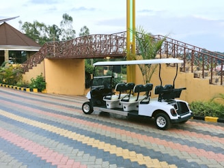 A white golf cart is parked on a multicolored brick-paved path. Behind it, there is a decorative bridge with intricate metal railings and stairs leading up to it. The area is landscaped with plants, and part of a building with a sloped roof is visible on the left. Yellow and black striped curb markings are present along the path.
