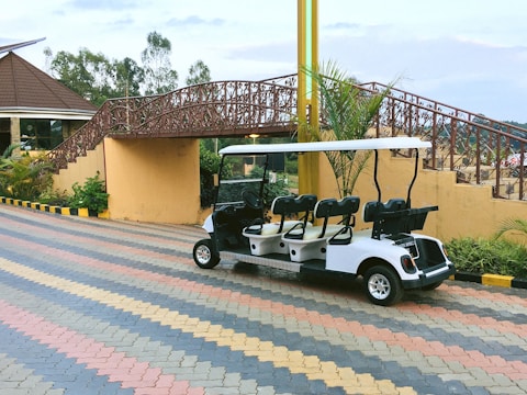 A white golf cart is parked on a multicolored brick-paved path. Behind it, there is a decorative bridge with intricate metal railings and stairs leading up to it. The area is landscaped with plants, and part of a building with a sloped roof is visible on the left. Yellow and black striped curb markings are present along the path.