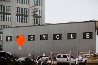 A large industrial building with a gray corrugated metal facade displays the word 'BICYCLE' in black windows with white letters. In the foreground, there is a road intersection with several cars and a bright orange road work sign. Power lines and a multi-story office building with many windows are visible in the background.