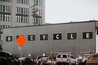 A large industrial building with a gray corrugated metal facade displays the word 'BICYCLE' in black windows with white letters. In the foreground, there is a road intersection with several cars and a bright orange road work sign. Power lines and a multi-story office building with many windows are visible in the background.