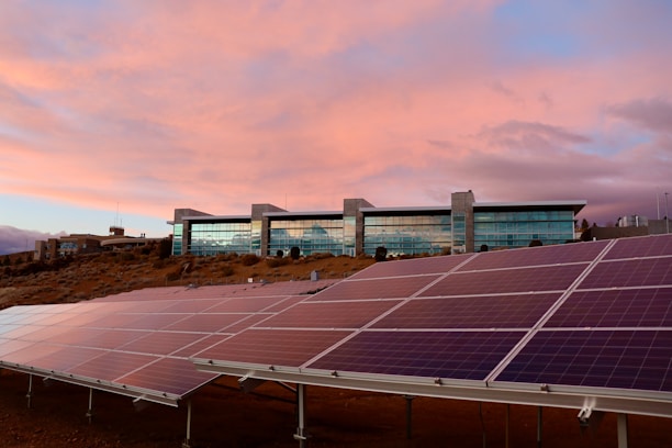 A modern building with large glass windows stands on a slight hill against a sky painted with soft pink and orange hues. In the foreground, well-aligned solar panels are installed on the ground, angled to capture sunlight.