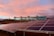 A modern building with large glass windows stands on a slight hill against a sky painted with soft pink and orange hues. In the foreground, well-aligned solar panels are installed on the ground, angled to capture sunlight.