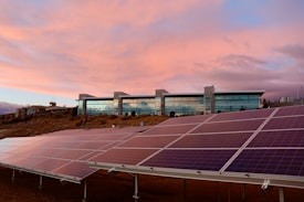 A modern building with large glass windows stands on a slight hill against a sky painted with soft pink and orange hues. In the foreground, well-aligned solar panels are installed on the ground, angled to capture sunlight.