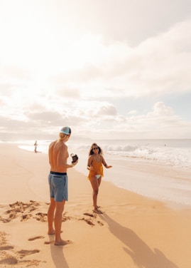 A sunny beach scene with a man and woman interacting playfully near the shoreline. The man wears shorts and a cap while holding a camera, and the woman is in a swimsuit and cover-up with sunglasses, smiling as she faces him. The sandy beach extends into the distance with gentle waves rolling in. There are a few other people visible further down the beach.