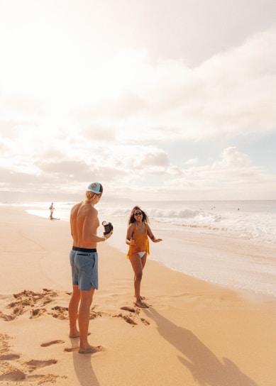 A sunny beach scene with a man and woman interacting playfully near the shoreline. The man wears shorts and a cap while holding a camera, and the woman is in a swimsuit and cover-up with sunglasses, smiling as she faces him. The sandy beach extends into the distance with gentle waves rolling in. There are a few other people visible further down the beach.