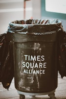 A large black trash can with plastic lining, labeled 'Times Square Alliance,' is situated on a street. The can appears to be well-used, with visible scuffs and marks on its surface.
