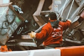 A person wearing a red jacket and gloves is working with a garbage truck, operating mechanisms to lift a dumpster filled with trash. The setting appears to be urban with a focus on waste management.