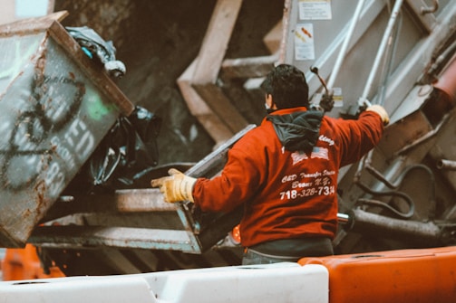 A person wearing a red jacket and gloves is working with a garbage truck, operating mechanisms to lift a dumpster filled with trash. The setting appears to be urban with a focus on waste management.