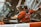 A person wearing a red jacket and gloves is working with a garbage truck, operating mechanisms to lift a dumpster filled with trash. The setting appears to be urban with a focus on waste management.