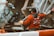 A person wearing a red jacket and gloves is working with a garbage truck, operating mechanisms to lift a dumpster filled with trash. The setting appears to be urban with a focus on waste management.
