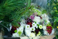 A collection of floral arrangements featuring white, red, and purple flowers mixed with greenery, including long pine needles. The bouquets are wrapped in clear plastic, highlighting the vibrant colors and diverse textures of the blooms.