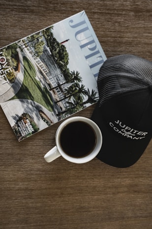 A magazine featuring an outdoor scene with a lighthouse and palm trees is placed alongside a white coffee cup filled with black coffee. Nearby, a black baseball cap with text is present. The background is a wooden surface.