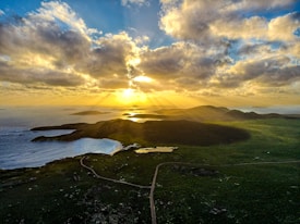 A scenic landscape featuring a coastline with waves gently lapping at a curved beach. The foreground is dominated by lush green fields, with a winding road leading towards distant hills. The sun is setting or rising on the horizon, casting dramatic golden rays through a partly cloudy sky, creating a radiant and serene atmosphere.