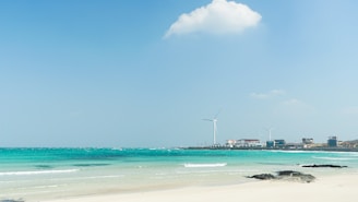 A serene beach scene with clear turquoise waters meeting a sandy shore. Wind turbines are visible in the background, near a small cluster of buildings. A single fluffy white cloud floats in the blue sky.