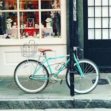 A turquoise bicycle is parked against a black post on a sidewalk in front of a store window. The window displays an array of jewelry items, including necklaces and bracelets, with a festive backdrop featuring snowflakes. The bicycle has a wire basket on the front and a brown saddle.