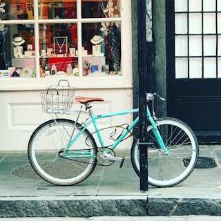 A turquoise bicycle is parked against a black post on a sidewalk in front of a store window. The window displays an array of jewelry items, including necklaces and bracelets, with a festive backdrop featuring snowflakes. The bicycle has a wire basket on the front and a brown saddle.
