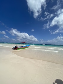 A vibrant beach scene features a small, colorful boat with a red canopy resting on the light sandy shore. The water displays a gradient of turquoise hues, and a deep blue sky is filled with scattered, fluffy white clouds. Other boats are visible in the distance, and a few people enjoy the water.