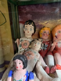 Several vintage or handmade dolls are displayed, each with distinct painted features and colorful clothing. The dolls have exaggerated facial expressions and are arranged closely together inside a glass cabinet.