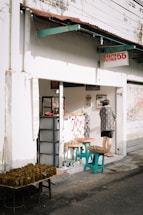 A small street-side shop with a sign that reads 'Bakpia Pathuk 55' and 'No 11'. The exterior is white with a rusty brown roof overhang. A person wearing a patterned shirt stands at the shop entrance. Several tables and stools outside the shop are displaying trays of food wrapped in banana leaves.