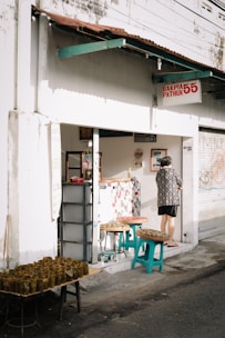 A small street-side shop with a sign that reads 'Bakpia Pathuk 55' and 'No 11'. The exterior is white with a rusty brown roof overhang. A person wearing a patterned shirt stands at the shop entrance. Several tables and stools outside the shop are displaying trays of food wrapped in banana leaves.