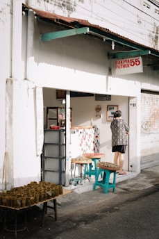 A small street-side shop with a sign that reads 'Bakpia Pathuk 55' and 'No 11'. The exterior is white with a rusty brown roof overhang. A person wearing a patterned shirt stands at the shop entrance. Several tables and stools outside the shop are displaying trays of food wrapped in banana leaves.