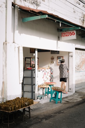 A small street-side shop with a sign that reads 'Bakpia Pathuk 55' and 'No 11'. The exterior is white with a rusty brown roof overhang. A person wearing a patterned shirt stands at the shop entrance. Several tables and stools outside the shop are displaying trays of food wrapped in banana leaves.