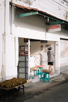A small street-side shop with a sign that reads 'Bakpia Pathuk 55' and 'No 11'. The exterior is white with a rusty brown roof overhang. A person wearing a patterned shirt stands at the shop entrance. Several tables and stools outside the shop are displaying trays of food wrapped in banana leaves.