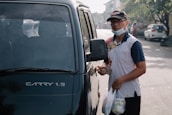 A person wearing a cap, glasses, and a mask below the chin stands beside a vehicle with a partially visible model name 'CARRY 1.5'. The person is holding a set of keys and two plastic bags containing food or goods. The scene appears to be on a street with a few cars and trees in the background, indicating an urban setting.