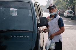 A person wearing a cap, glasses, and a mask below the chin stands beside a vehicle with a partially visible model name 'CARRY 1.5'. The person is holding a set of keys and two plastic bags containing food or goods. The scene appears to be on a street with a few cars and trees in the background, indicating an urban setting.