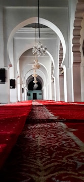 An interior view of a mosque corridor featuring elegant architectural arches. Ornate chandeliers hang from the ceiling, casting a warm glow on the surroundings. The floor is covered with a richly patterned red carpet extending into the distance.